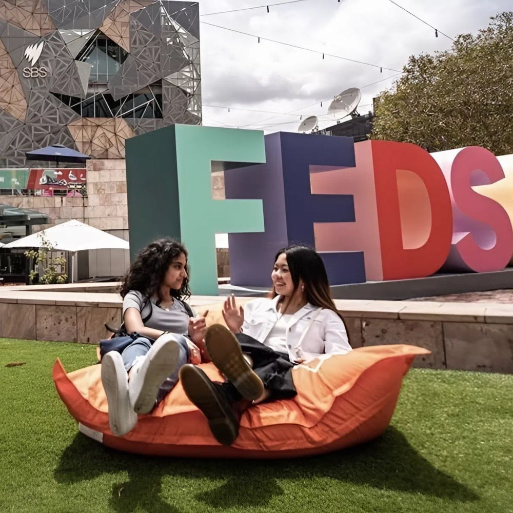 Two people relaxing on an orange Fed Square bean bag in front of the giant FEDSQ letters in Melbourne