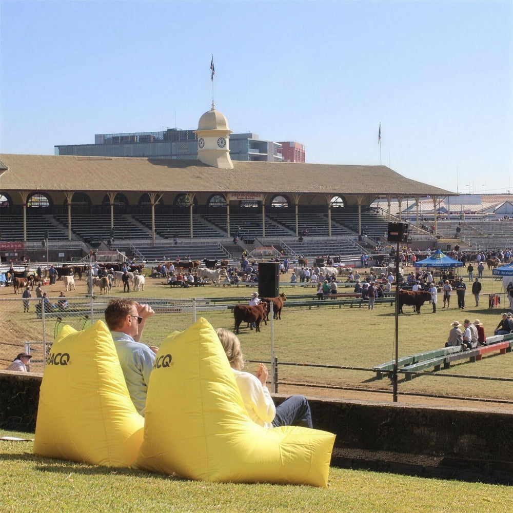 Two people relaxing on yellow RACQ bean bags beside the main arena during cattle judging at the Ekka, Brisbane