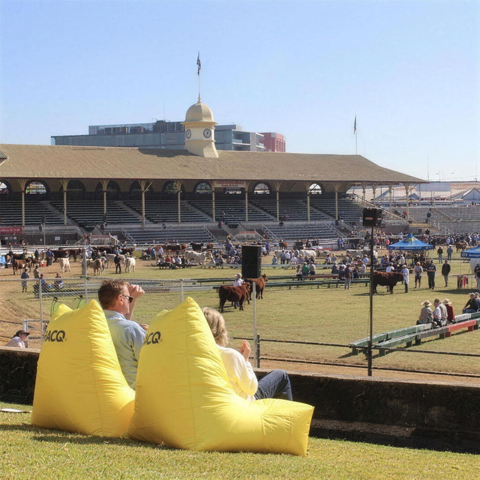 Two people relaxing on yellow RACQ bean bags beside the main arena during cattle judging at the Ekka, Brisbane