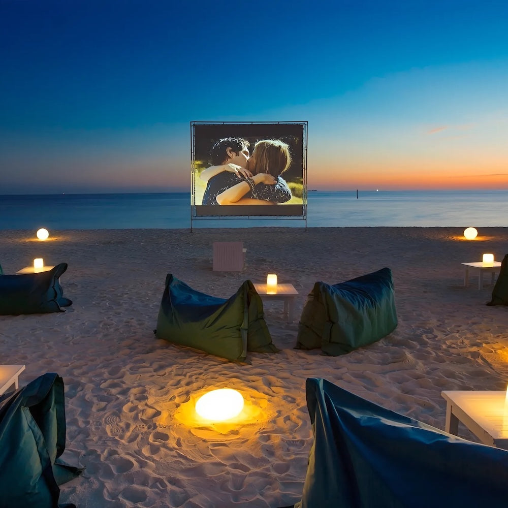Beachfront outdoor cinema at dusk at Velassaru Maldives, with Hayman bean bags on the sand facing the screen and the Indian Ocean behind