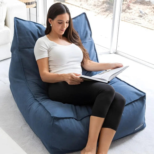 Woman reading a book on a large blue bean bag chair in a bright room.