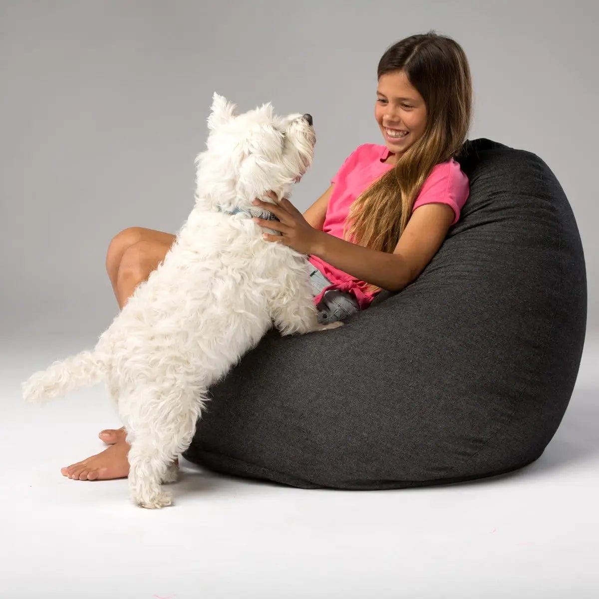 Woman sitting on a dark gray bean bag with a white dog, both smiling.