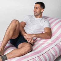 Man sitting on a pink and white striped bed wearing a white t-shirt and dark shorts.