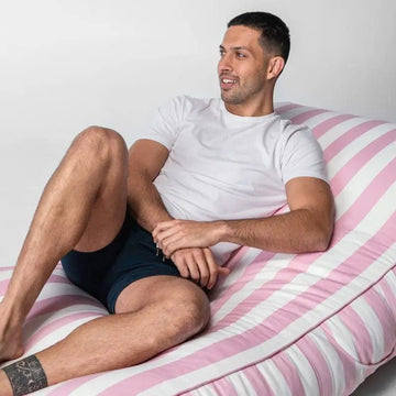 Man sitting on a pink and white striped bed wearing a white t-shirt and dark shorts.